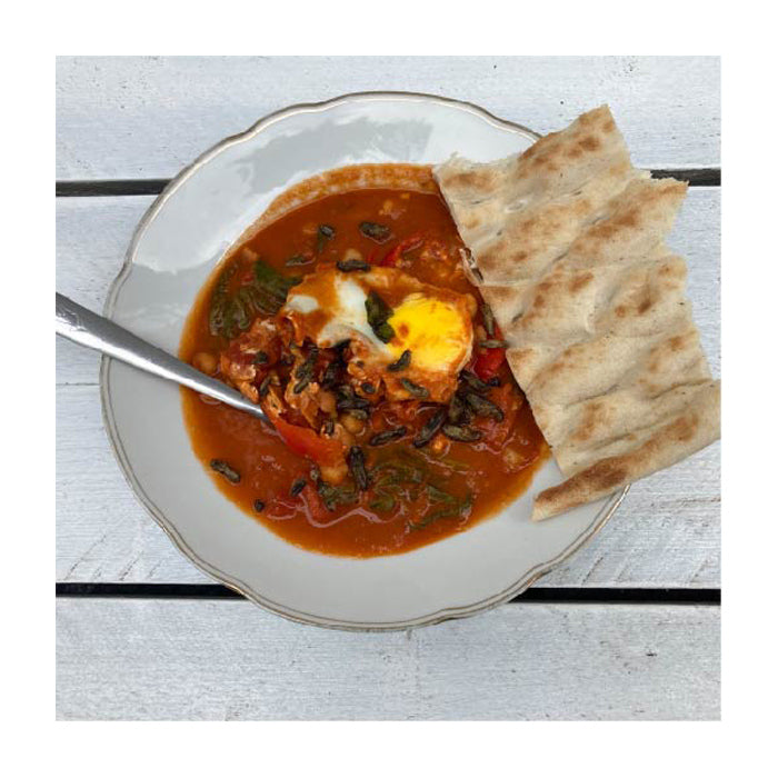 Shakshuka angerichtet auf einem weißen Teller, welcher auf einem weißen Holztisch steht. In dem Rot der Tomatensoße sind ein Ei und geröstete Grillen zu erkennen. Auf dem Tellerrand liegt ein Stück helles Fladenbrot.