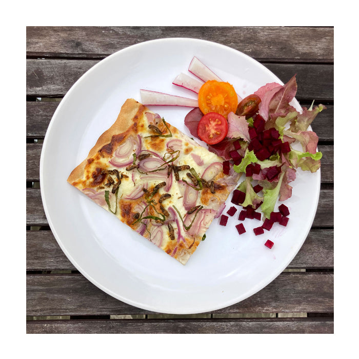 Ein Stück Flammkuchen auf weißem Teller, angerichtet mit Blattsalat, roter Beete und Tomatenscheiben. Der Teller steht auf einem Holztisch.