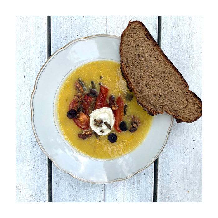 Ein Teller Zucchinisuppe auf weißem Holztisch, angerichtet mit Tomaten, Oliven und Walnüssen. Auf dem Tellerrand liegt eine Scheibe Sauerteigbrot. 