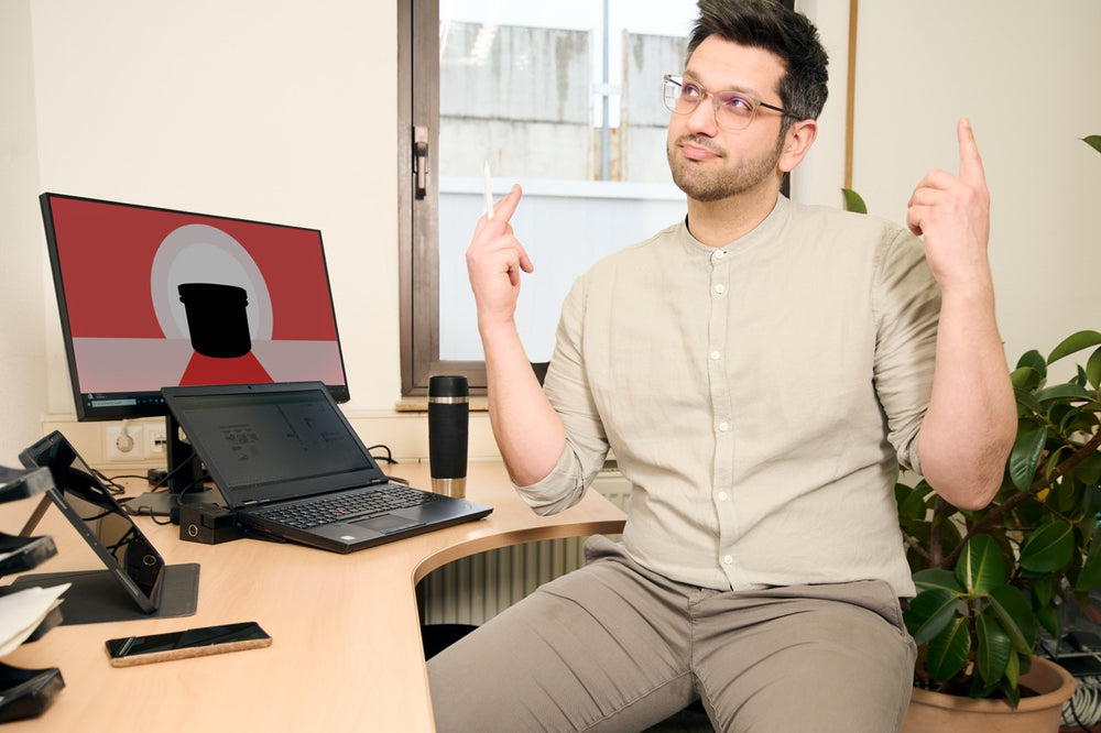 Foto eines Mitarbeiters an einem Bürotisch. Auf dem Tisch stehen Tablet, Laptop und ein Monitor, auf dem eine bunte Illustration zu sehen ist.
